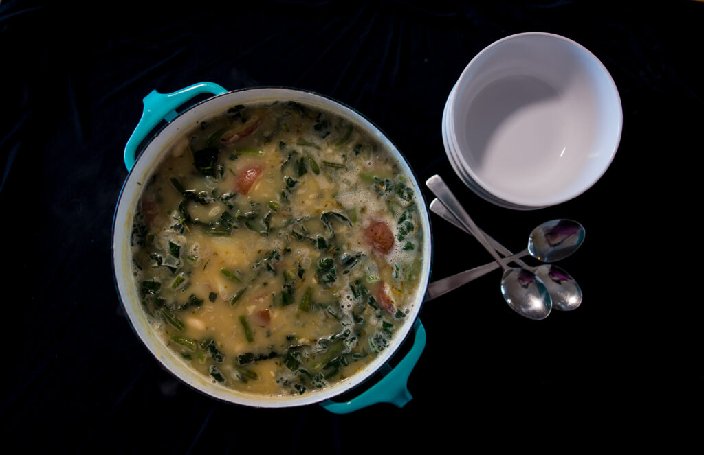 Kale and potato soup in blue dansk pot with stack of spoons on the side on a black background
