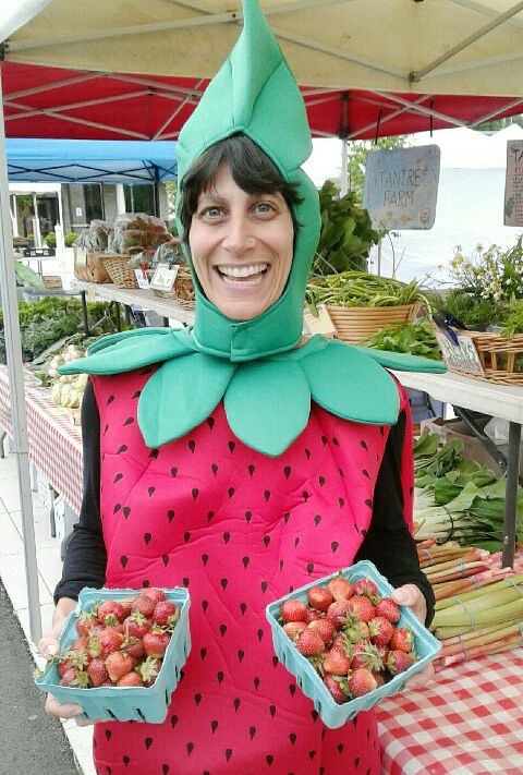 A woman is dressed in a strawberry costume, holding strawberries, at the Chelsea Farmers Market.