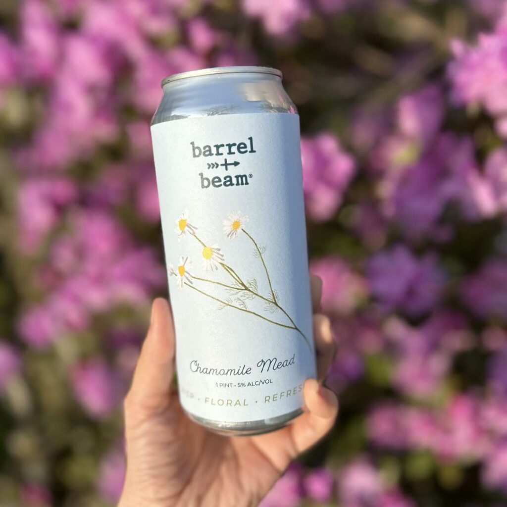 A hand holds up Barrel + Beam's chamomile mead can in front of a background of purple flowers. Photo credit: Barrel + Beam