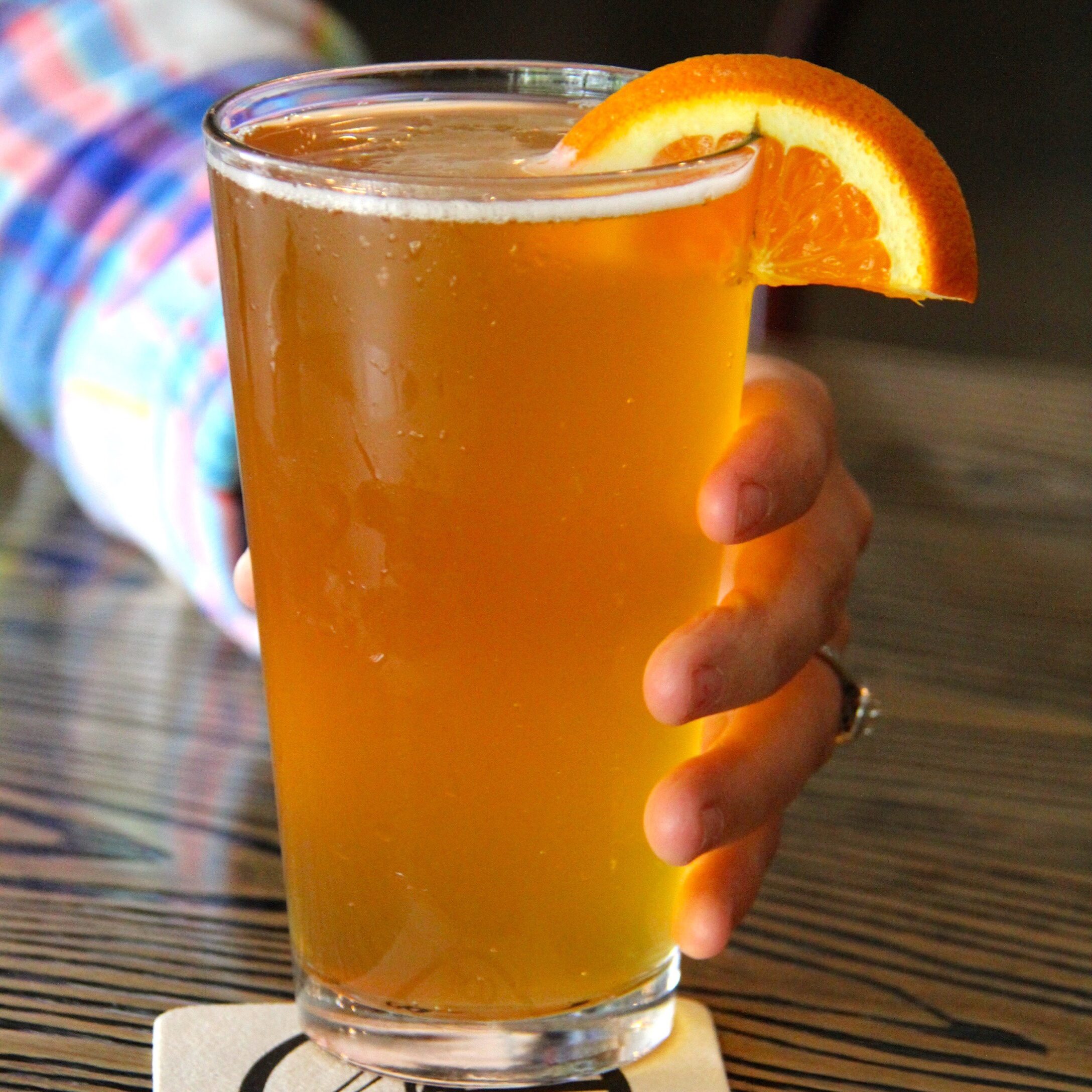 A person in a checked shirt with a ring holds an amber beer with an orange slice as a garnish. Photo credit: Les Cheneaux Distillers