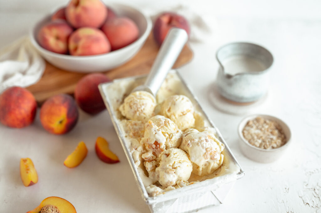 Peach Crumble ice cream in a loaf pan with multiple scoops, with fresh peaches scattered around. Photo Credit: Taste the Local Difference 