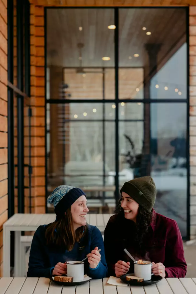 Two friends in warm hats smile at each other while enjoying soup in a mug at Farm Club. Photo credit: Courtney Kent 